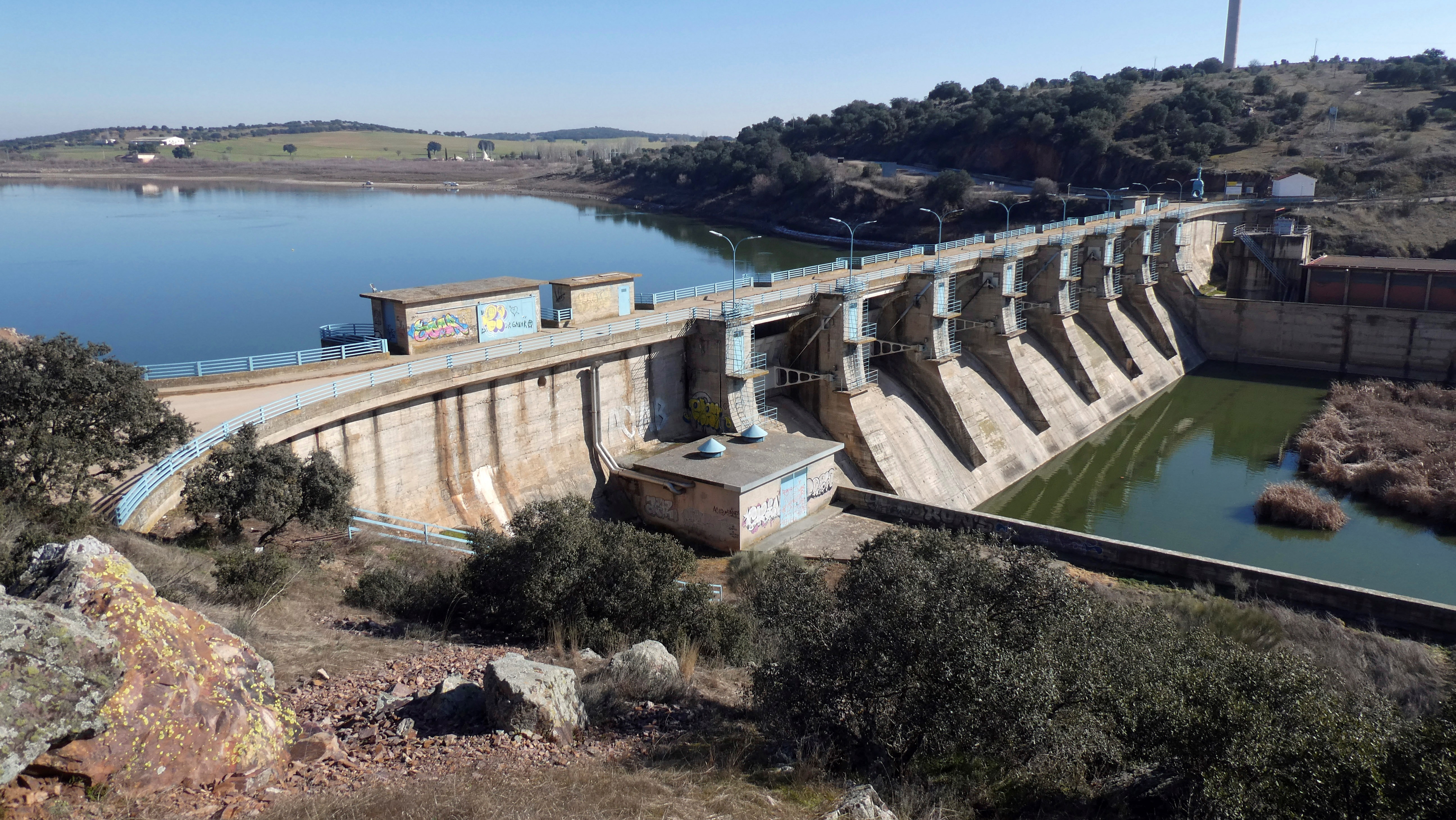 Presa de hormigón con compuertas metálicas sobre un río, rodeada de vegetación y colinas, con el embalse de agua al fondo bajo cielo despejado.