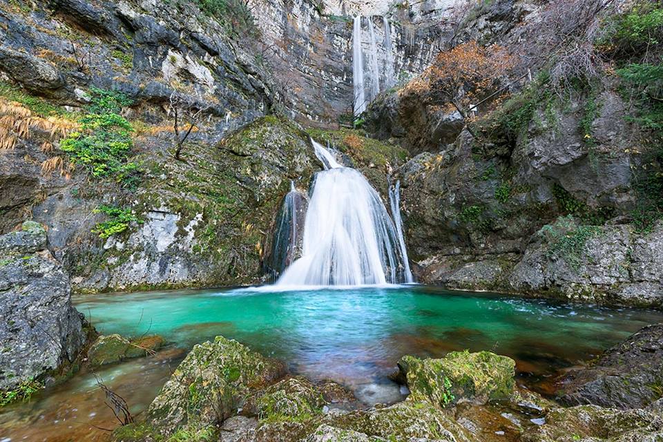 Cascada del río Mundo cayendo entre rocas hacia una poza de agua turquesa, rodeada de vegetación y paredes rocosas en un entorno natural.